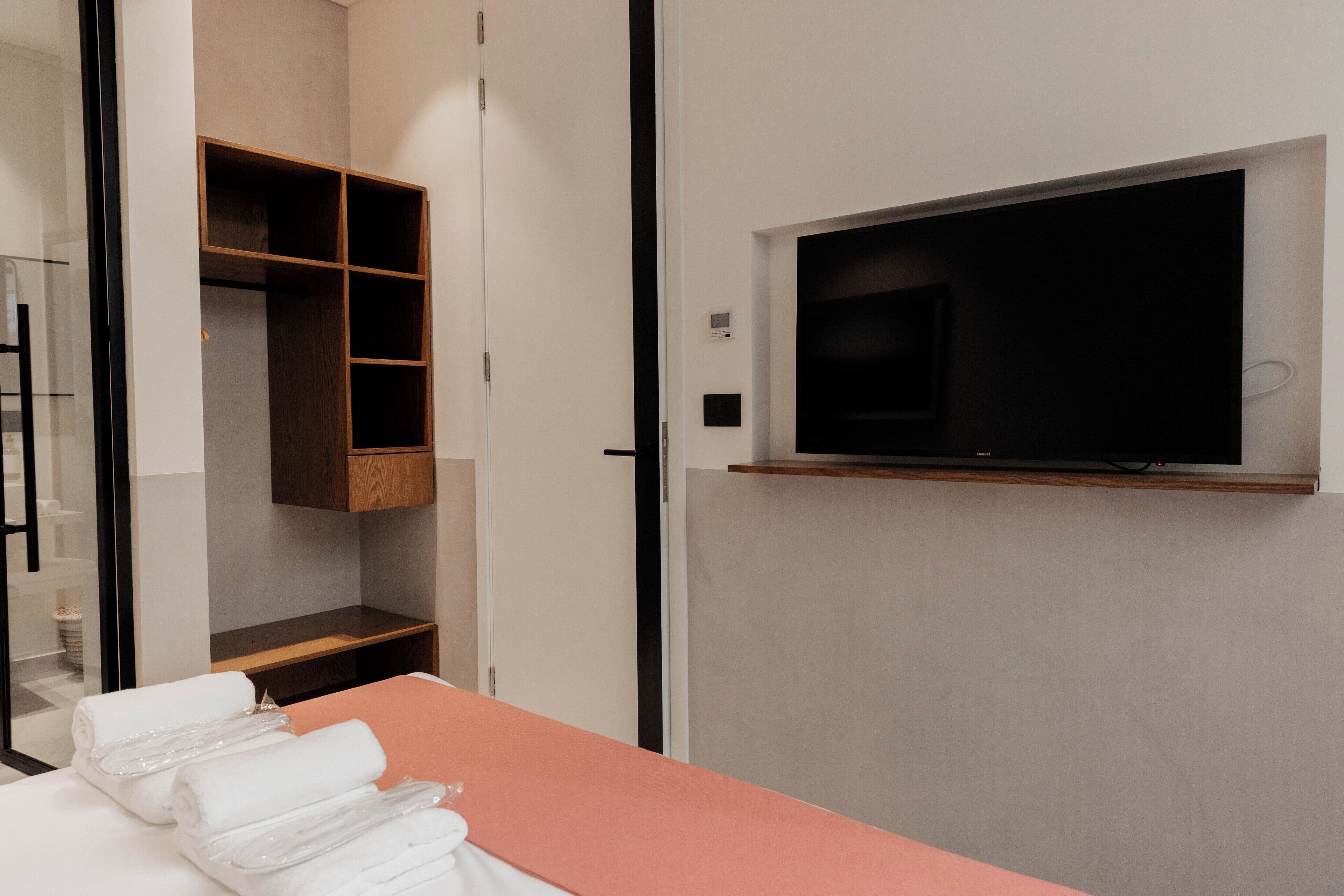 Una habitación de hotel con cama, televisión montada en la pared, armario de madera y zona de ducha con mampara de cristal.