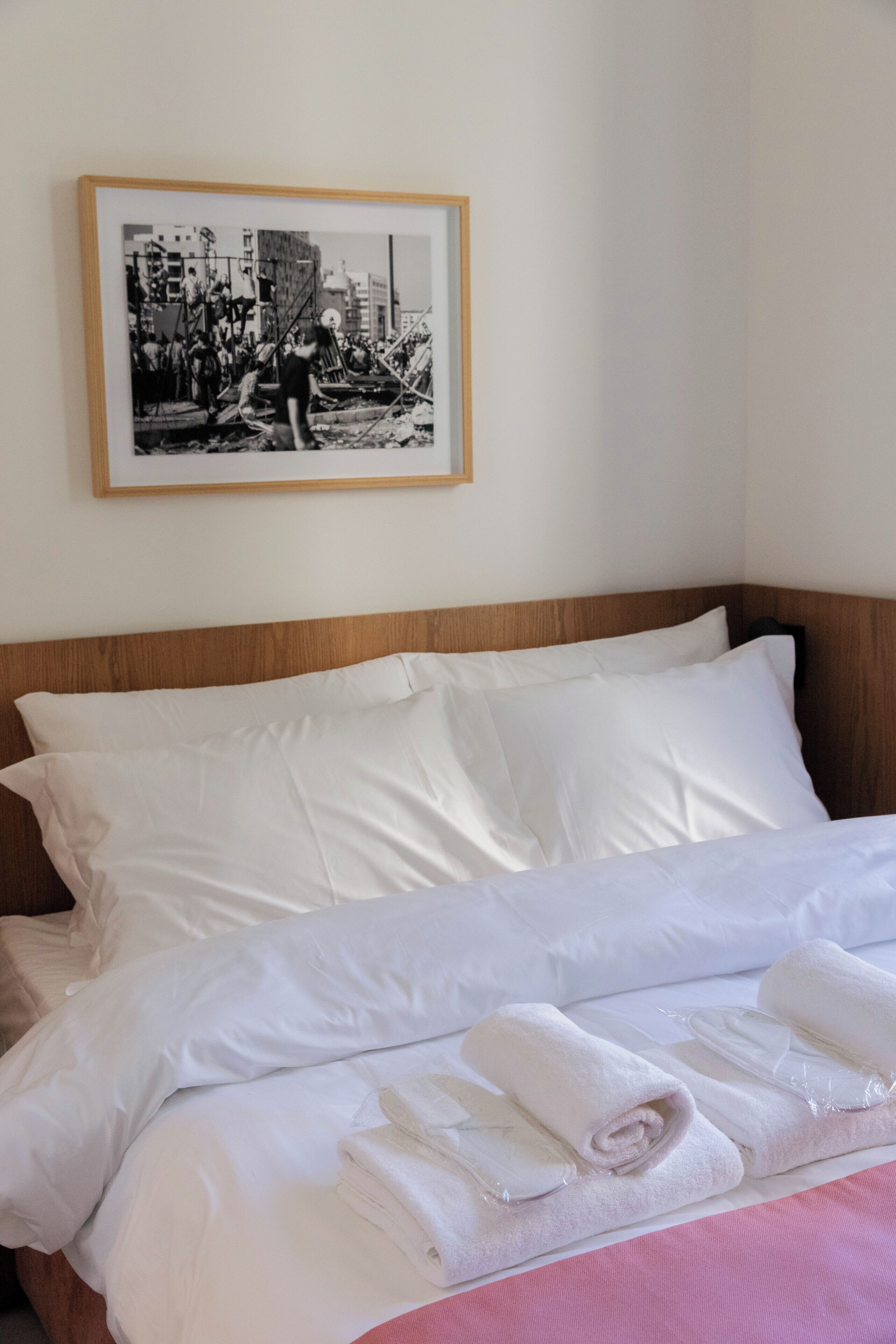 A neatly made bed with white linens and rolled towels, a framed black and white photo on the wall, and a wooden headboard.