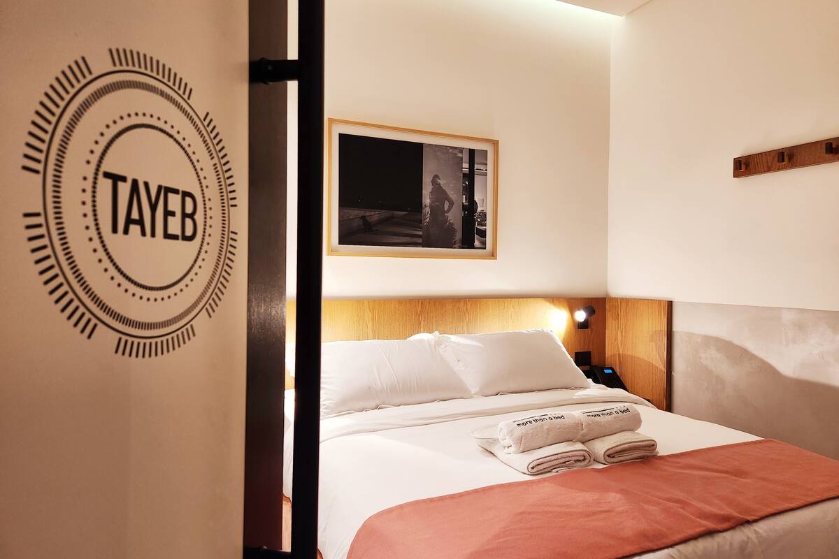Una habitación de hotel con una cama, una mesita de noche, una lámpara, un cuadro enmarcado en la pared y una puerta con un logo circular.