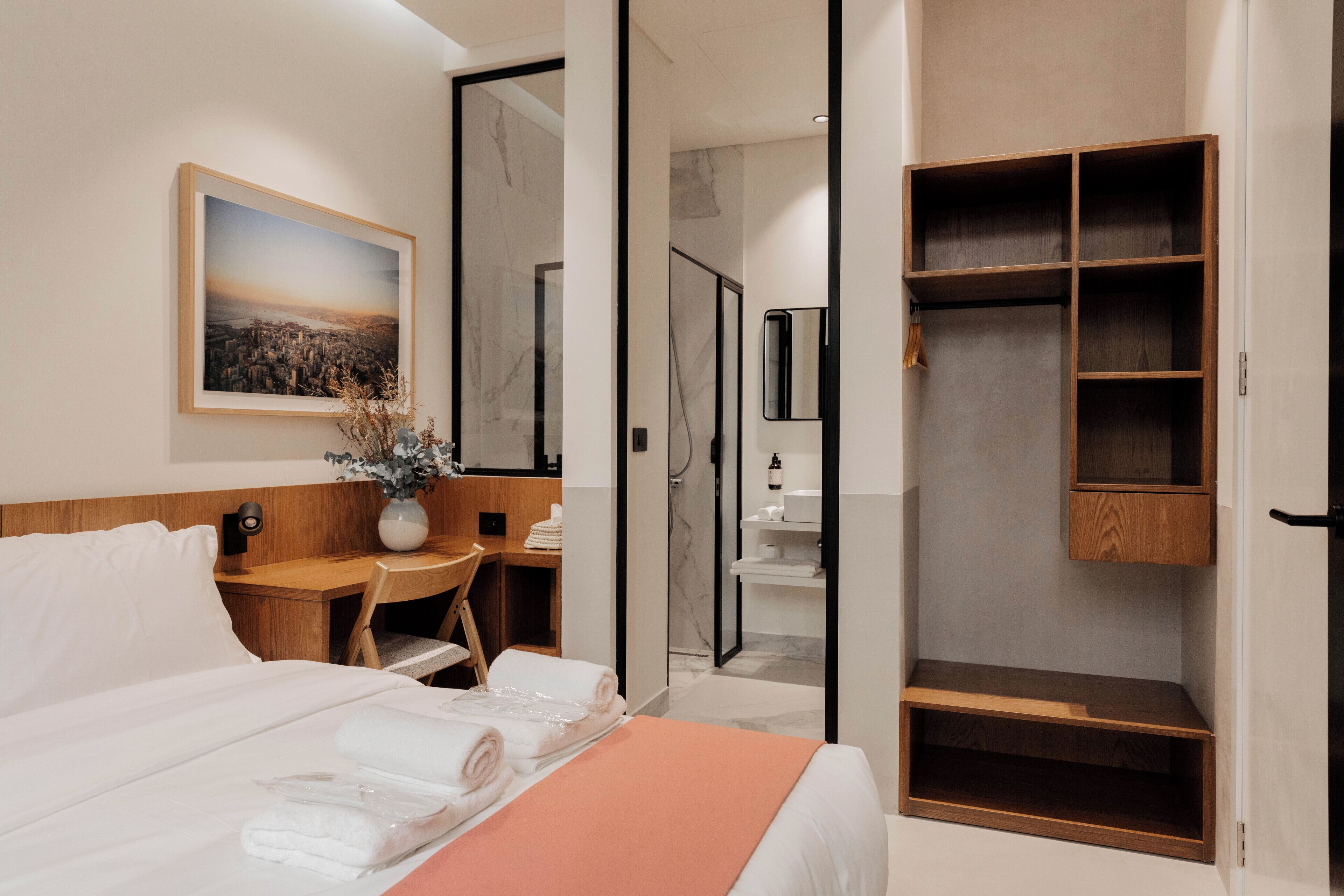 A modern hotel room with a neatly made bed, a desk with a vase of flowers, a walk-in closet, and a bathroom with a shower and mirror.