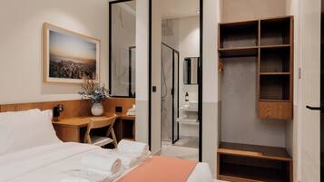 A modern hotel room with a neatly made bed, a desk with a vase of flowers, a walk-in closet, and a bathroom with a shower and mirror.