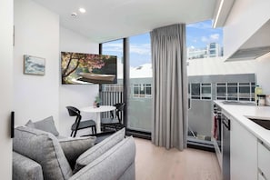Un salon moderne avec un canapé gris, une table à manger avec des chaises, un tableau et une grande fenêtre donnant sur des immeubles.