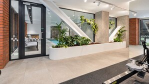 A modern indoor space with a glass door entrance, a seating area with a sofa and coffee table, a large planter with greenery, and a rowing machine. - Hyde Lane 1BR | Walk to Cafes + Pool + Gym (Wellington)