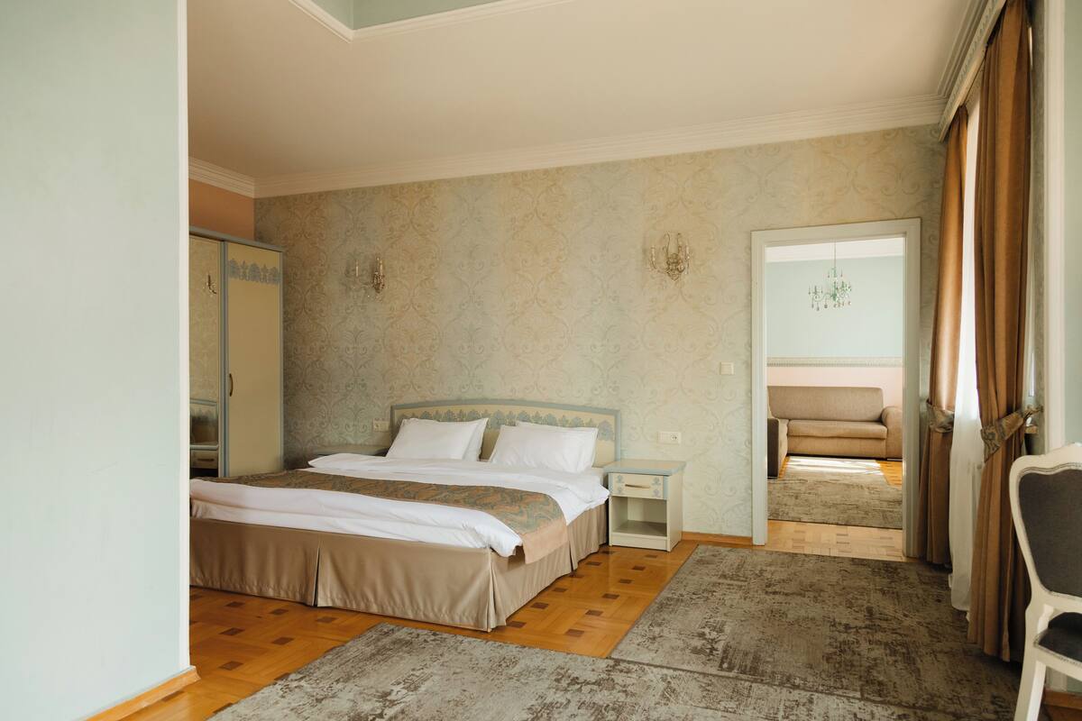 Une chambre avec un grand lit, une table de chevet, une armoire et une vue sur le salon par une porte ouverte.