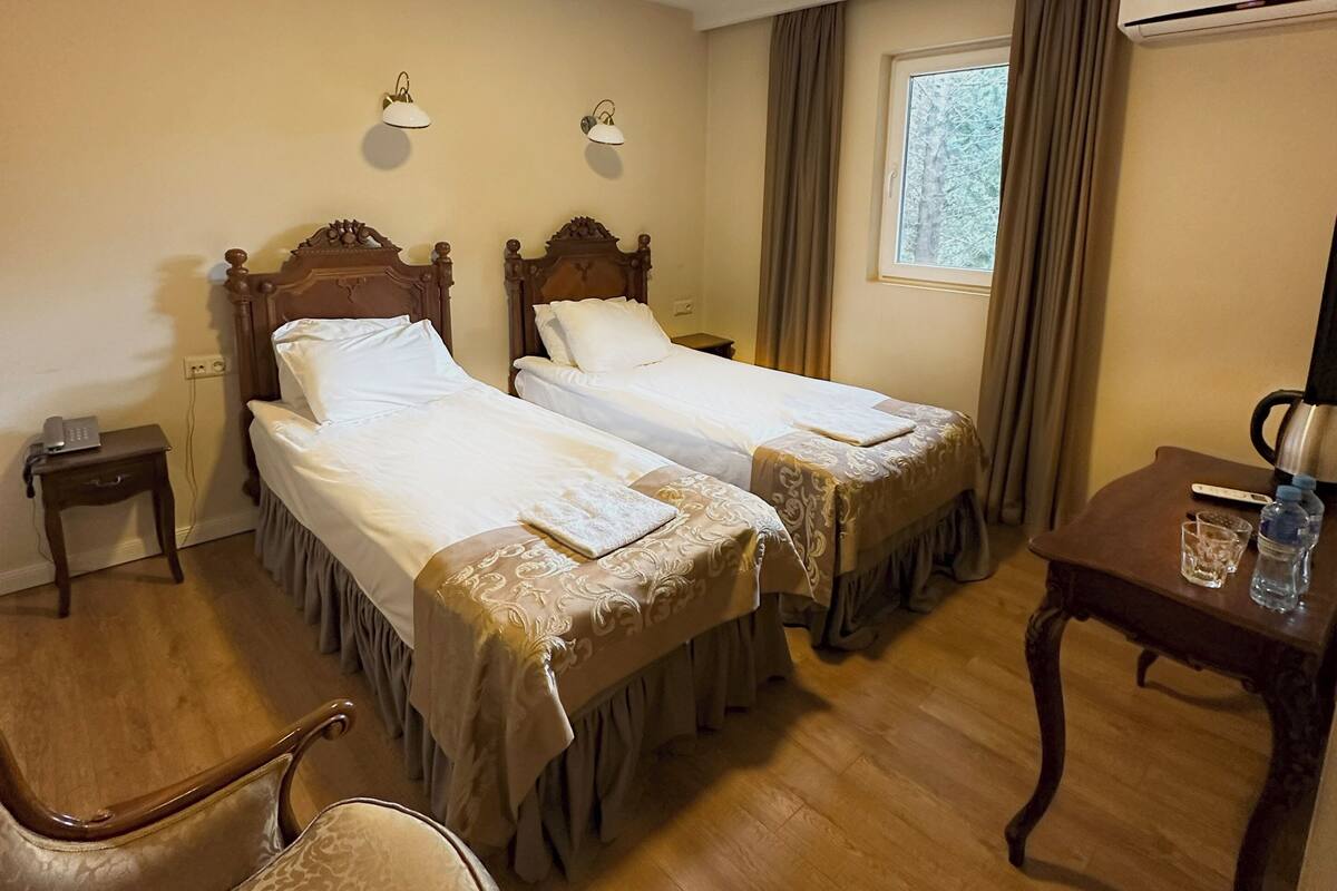Une chambre d’hôtel avec deux lits en bois, une petite table avec une bouteille d’eau, une chaise, une table de chevet avec une lampe et un appareil de climatisation.