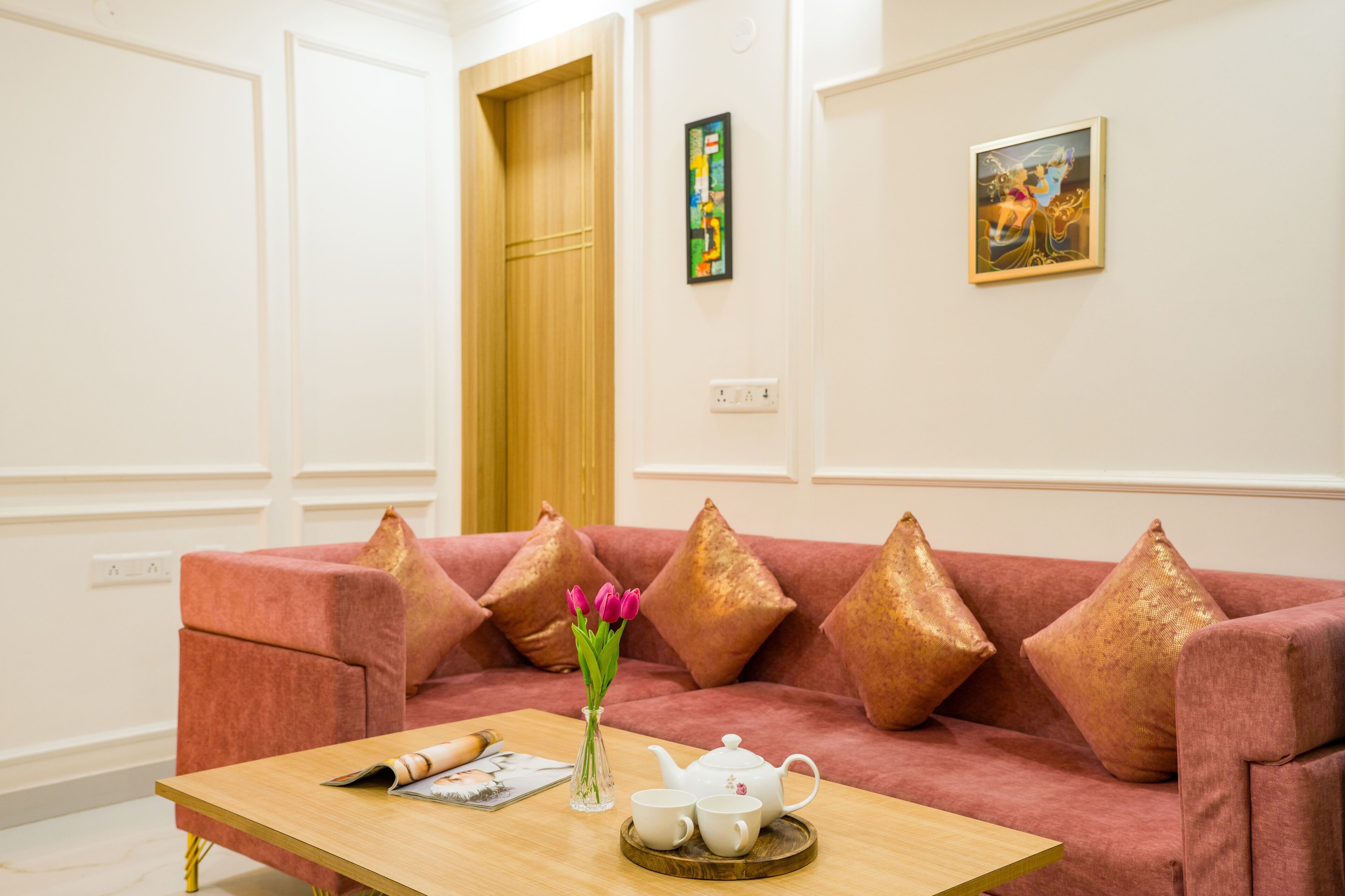A modern living room with a pink sofa, gold cushions, a wooden coffee table, a vase with flowers, and a teapot.