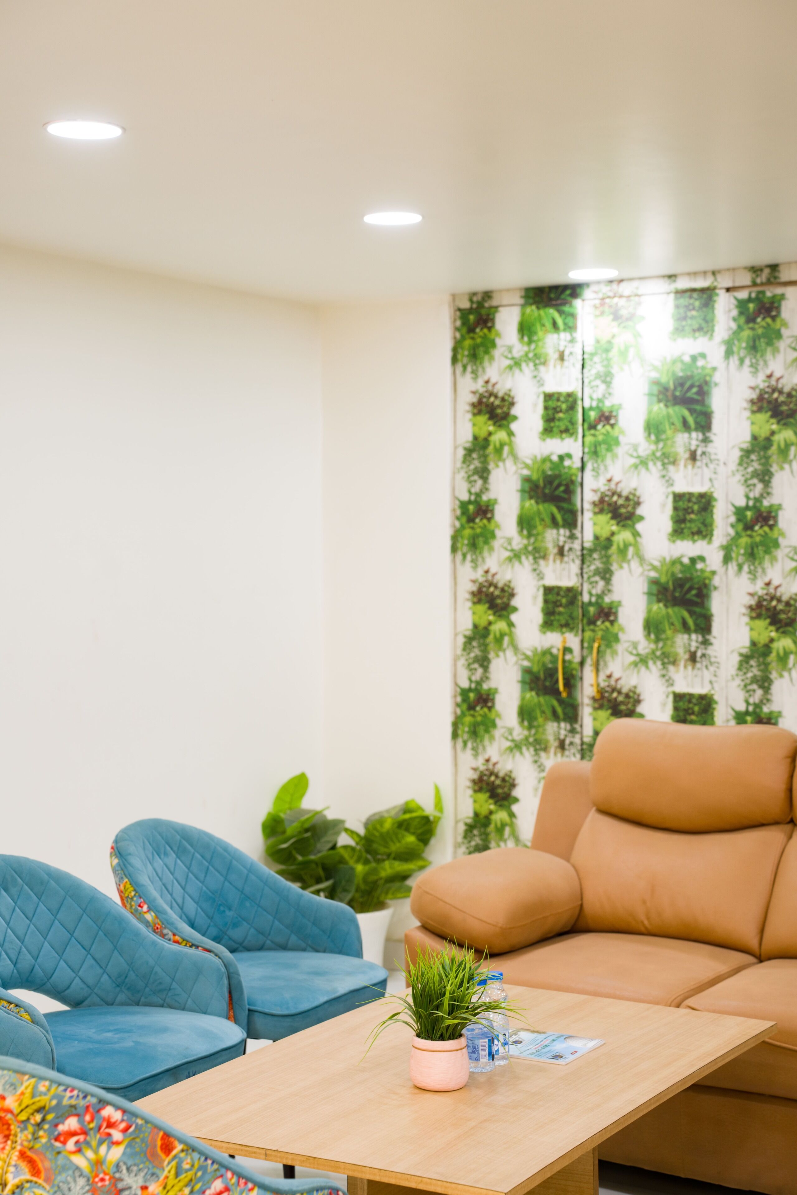 Una habitación con un sofá color beige, un sillón azul, una mesa de centro de madera con una planta y una botella, y una pared con estampado de plantas verdes.