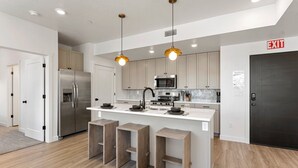 A modern kitchen with stainless steel appliances, wooden stools, and pendant lighting. - The Way Station 2 Bedroom Condo (Hurricane)