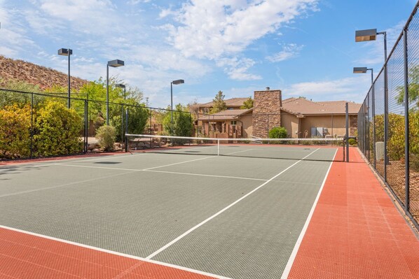 An outdoor tennis court with a net, surrounded by a fence, and adjacent to a building with a stone facade.