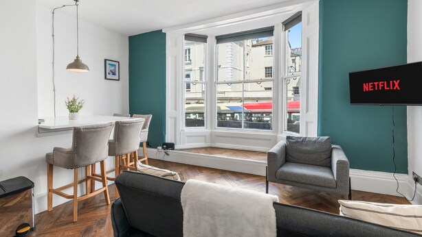 A modern living room with a dining area, a large window, and a TV mounted on the wall.