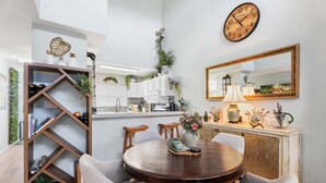 A dining area with a round wooden table, four chairs, a bookshelf, a kitchen in the background, and a wall clock. - Big Island Waikoloa Hills 606 2 Bedroom Condo (Waikoloa)