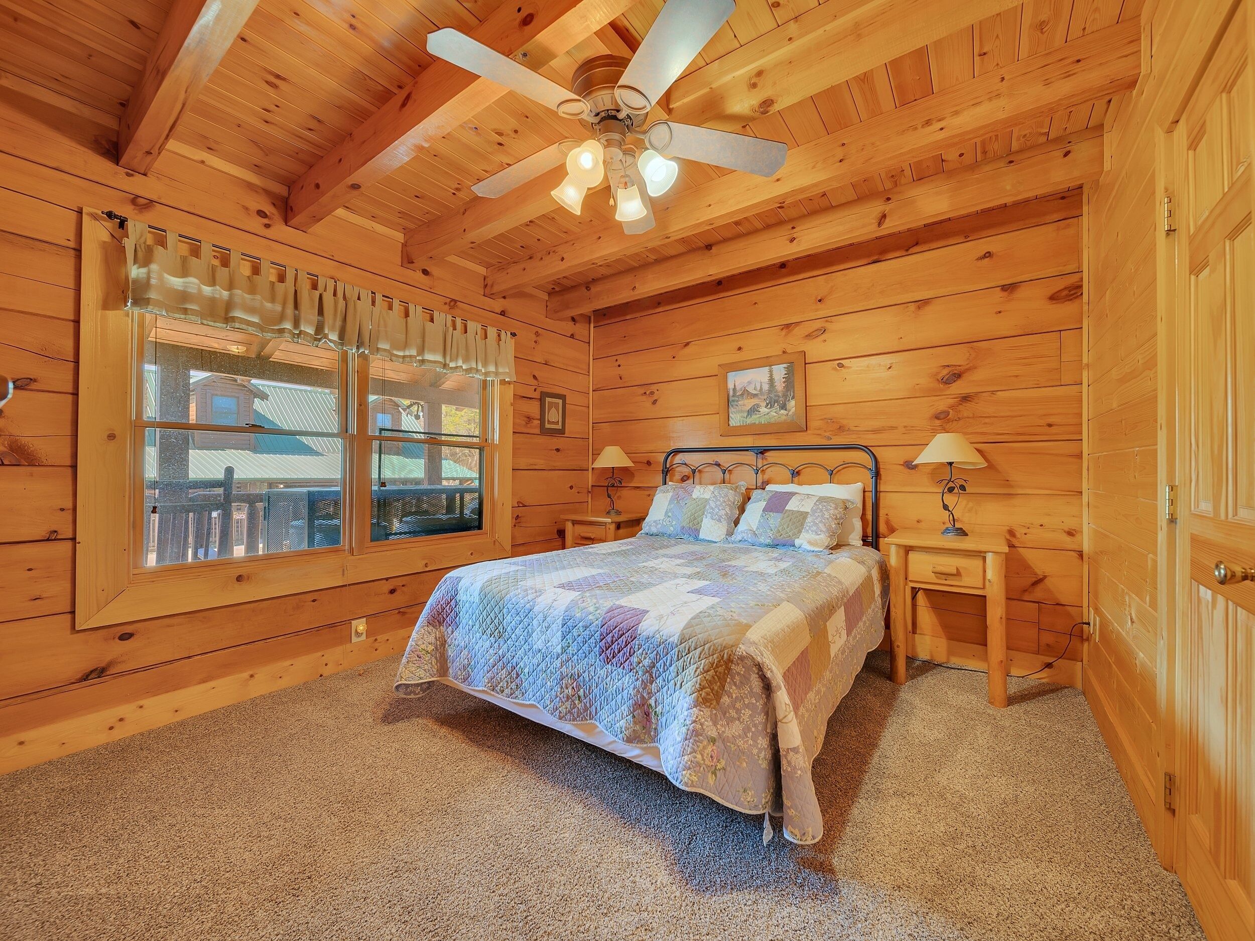 Ein Schlafzimmer in einer Holzhütte mit einem Bett, zwei Nachttischen, einem Deckenventilator und einem Fenster mit Vorhängen.