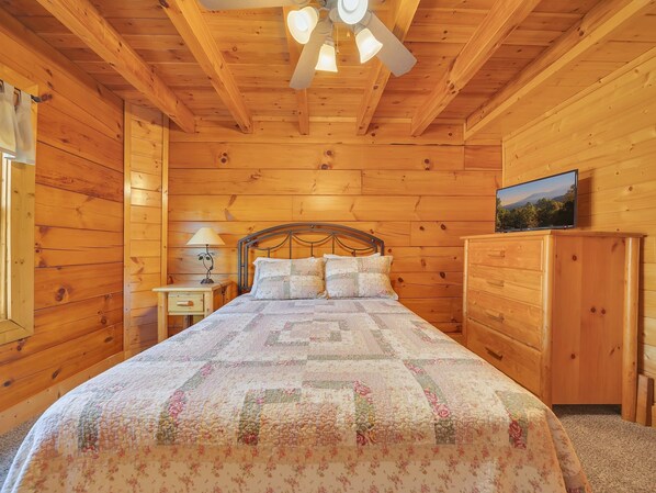 A wooden cabin bedroom with a bed, nightstand, lamp, dresser, and ceiling fan.