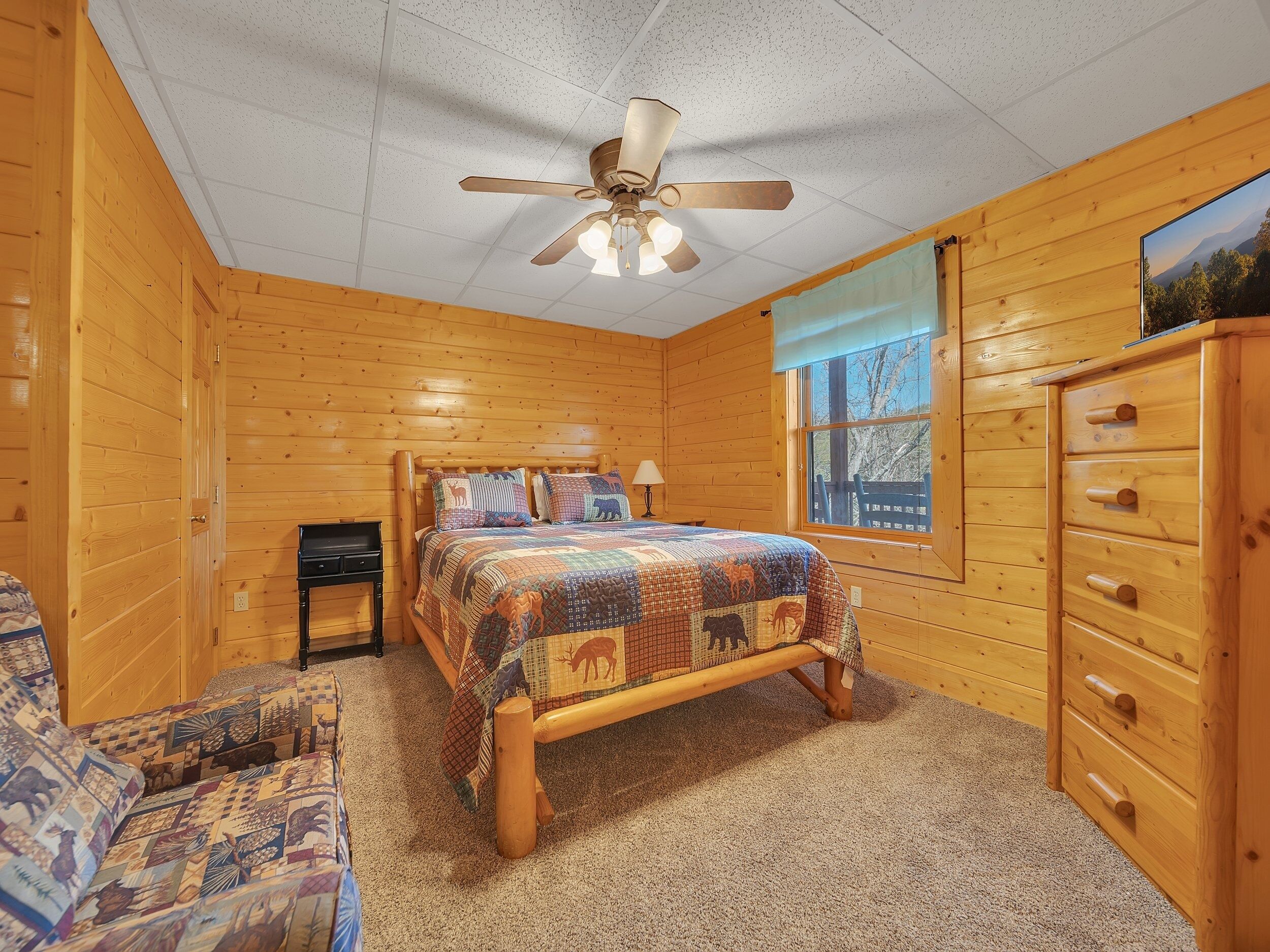 A wooden cabin bedroom with a bed, a dresser, a nightstand, and a window with a view of trees.
