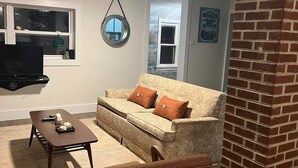 A living room with a sofa, coffee table, and a chair. There is a television, a ceiling fan, and a window with a view.