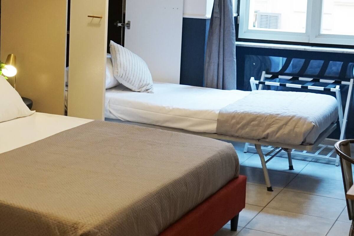 A hotel room with a bed, a bedside table, a lamp, a window with curtains, and a view of the outside.
