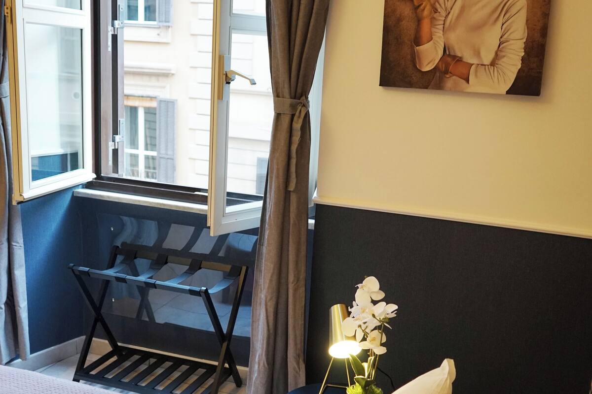 A hotel room with a bed, a desk with a chair, a window with curtains, and a painting on the wall.