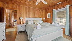Une chambre à coucher aux murs lambrissés, avec un lit blanc, deux tables de chevet, un ventilateur de plafond et une fenêtre avec des rideaux.