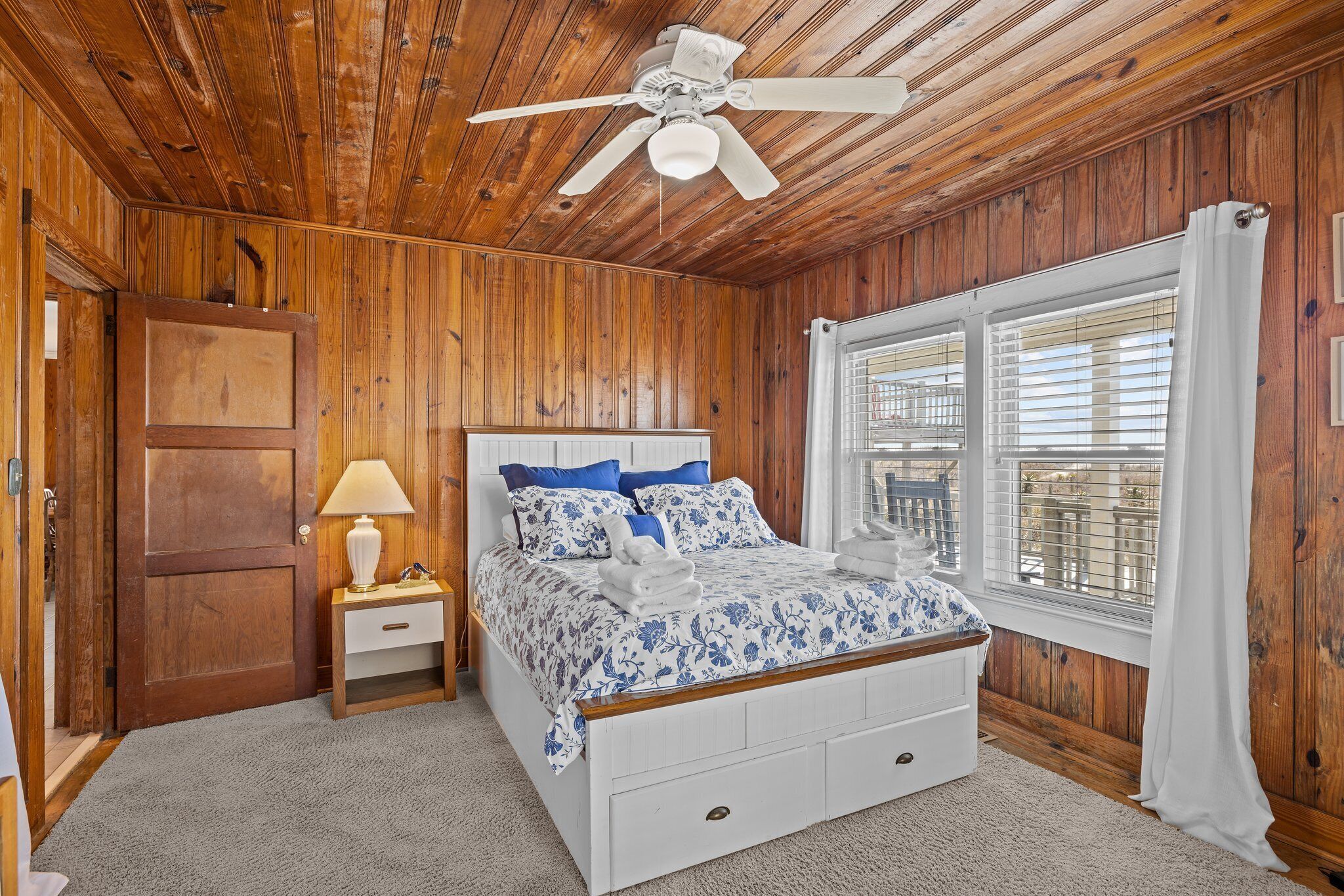 A bedroom with a wooden ceiling, a ceiling fan, a bed with blue floral bedding, a nightstand with a lamp, and a window with blinds.
