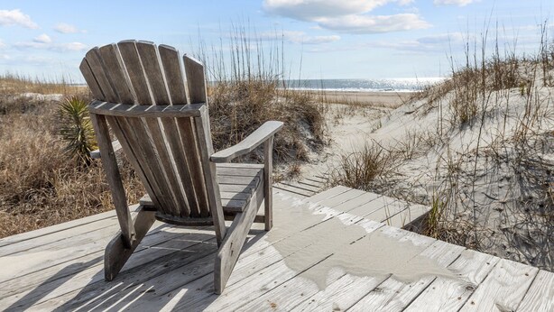 Une chaise Adirondack en bois est posée sur une terrasse en bois, d’où l’on a vue sur une plage, des dunes de sable et l’océan en arrière-plan.