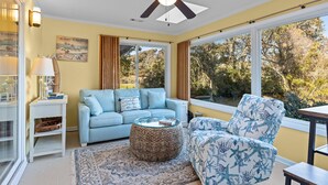 Ein Wohnzimmer mit Deckenventilator, einem blauen Sofa, einem gemusterten Sessel und großen Fenstern mit Blick auf Bäume.