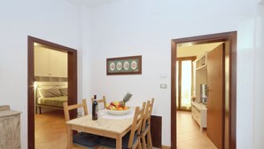 A dining area with a wooden table, chairs, and a fruit bowl. There is a bed in the adjacent room and a white cabinet. - Trasimeno Holiday Home - Apartment Il Girasole (Castiglione del Lago)