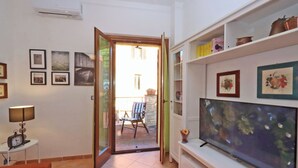 A room with a television, a table with a lamp, framed pictures on the wall, and a door leading to an outdoor area with a chair and table. - Trasimeno Holiday Home - Apartment Il Girasole (Castiglione del Lago)