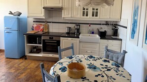 A compact kitchen with white cabinets, a blue refrigerator, a dining table with a floral tablecloth, and a hanging light fixture. - "Himmelsnest" at Hof Wiesentau - Life in a thatched-roof house, 65 m², sleeps 4–5 (Bösdorf)