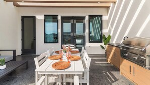 A modern outdoor dining area with a white table set for four, surrounded by white chairs, a black door, and a barbecue grill.