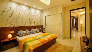 A hotel room with a marble-patterned wall, a large bed with a colorful bedspread, a nightstand with a lamp, and a wardrobe. - Premium Service Flats (Kharar)