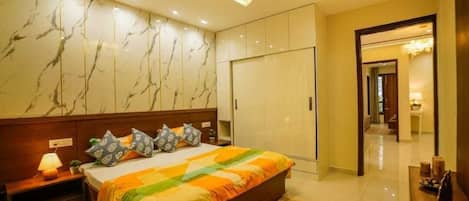 A hotel room with a marble-patterned wall, a large bed with a colorful bedspread, a nightstand with a lamp, and a wardrobe.