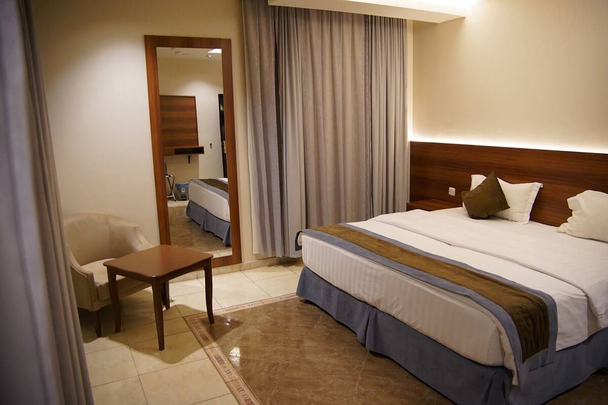 A hotel room with a bed, a chair, a small table, a mirror, and a view of the hallway.