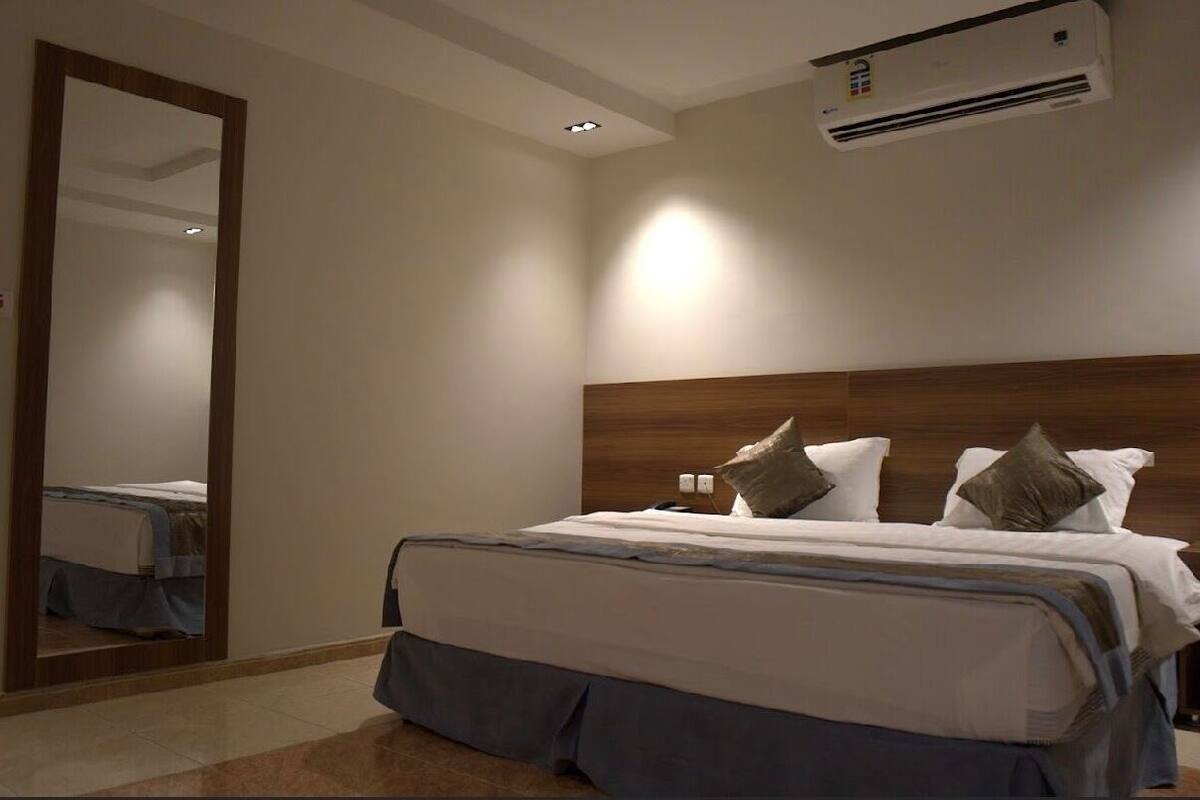 A bedroom with a large bed, a wooden headboard, a wall-mounted air conditioner, and a mirror reflecting part of the room.