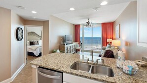 A hotel room with a kitchen area featuring granite countertops, a double sink, and a view of the ocean through a large window with red curtains.