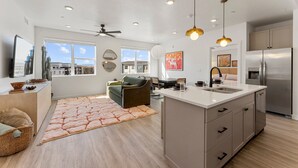 A modern kitchen with a central island, a dining area with a green sofa, and a large window offering a view of buildings. - The Way Station: Resort Pools, Lazy River, Waterslide + Pickleball (Hurricane)