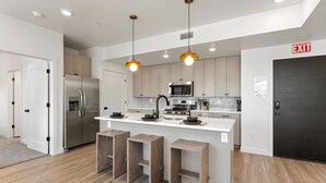 A modern kitchen with stainless steel appliances, wooden stools, and pendant lighting. - The Way Station: Resort Pools, Lazy River, Waterslide + Pickleball (Hurricane)