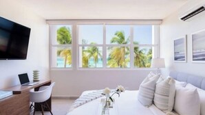 Een moderne slaapkamer met een groot bed, een bureau met een laptop, een televisie en een raam met uitzicht op palmbomen en de zee.
