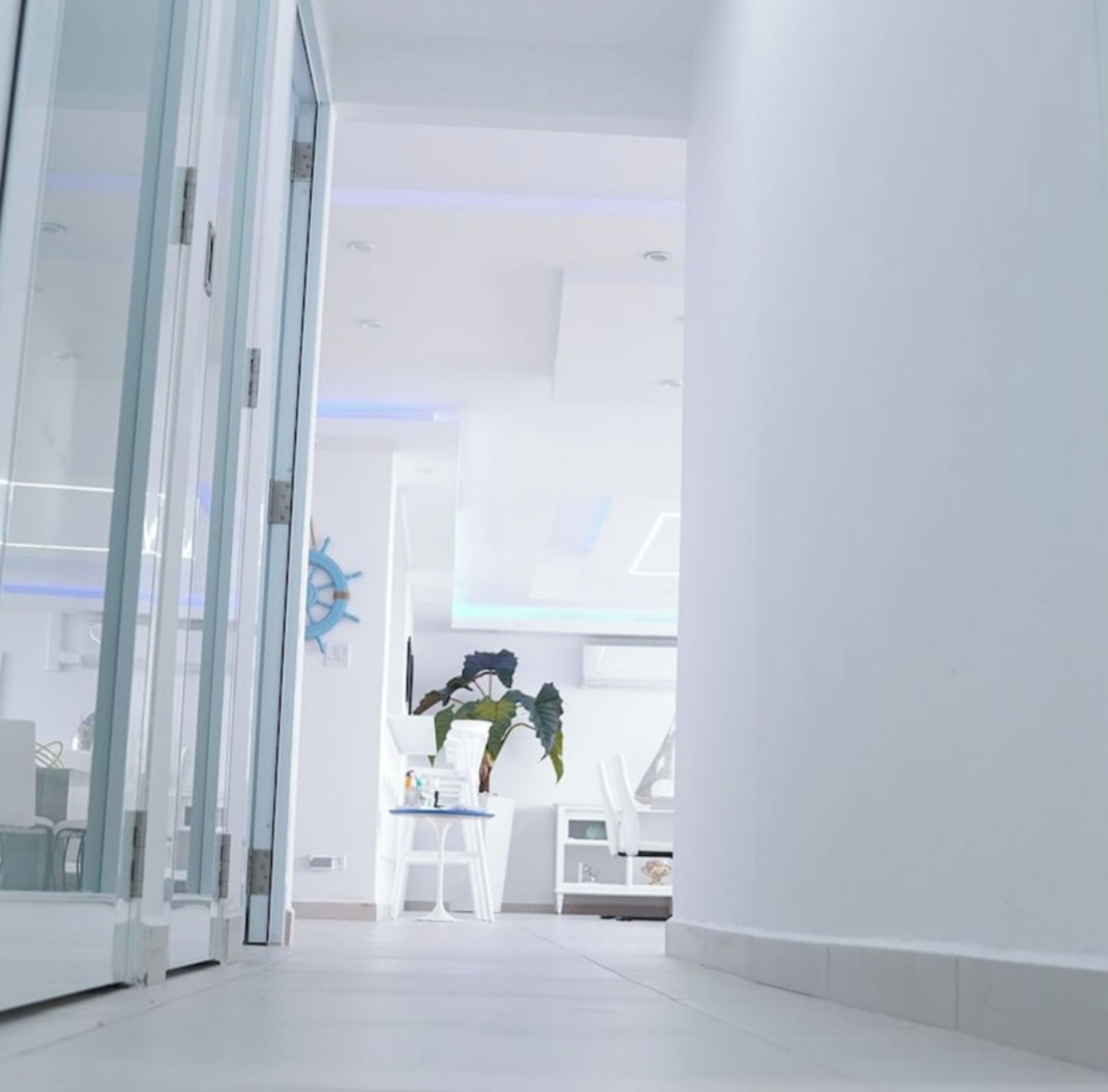 A modern, well-lit hallway with glass doors leading to a room. There is a plant and a small table with chairs in the room.