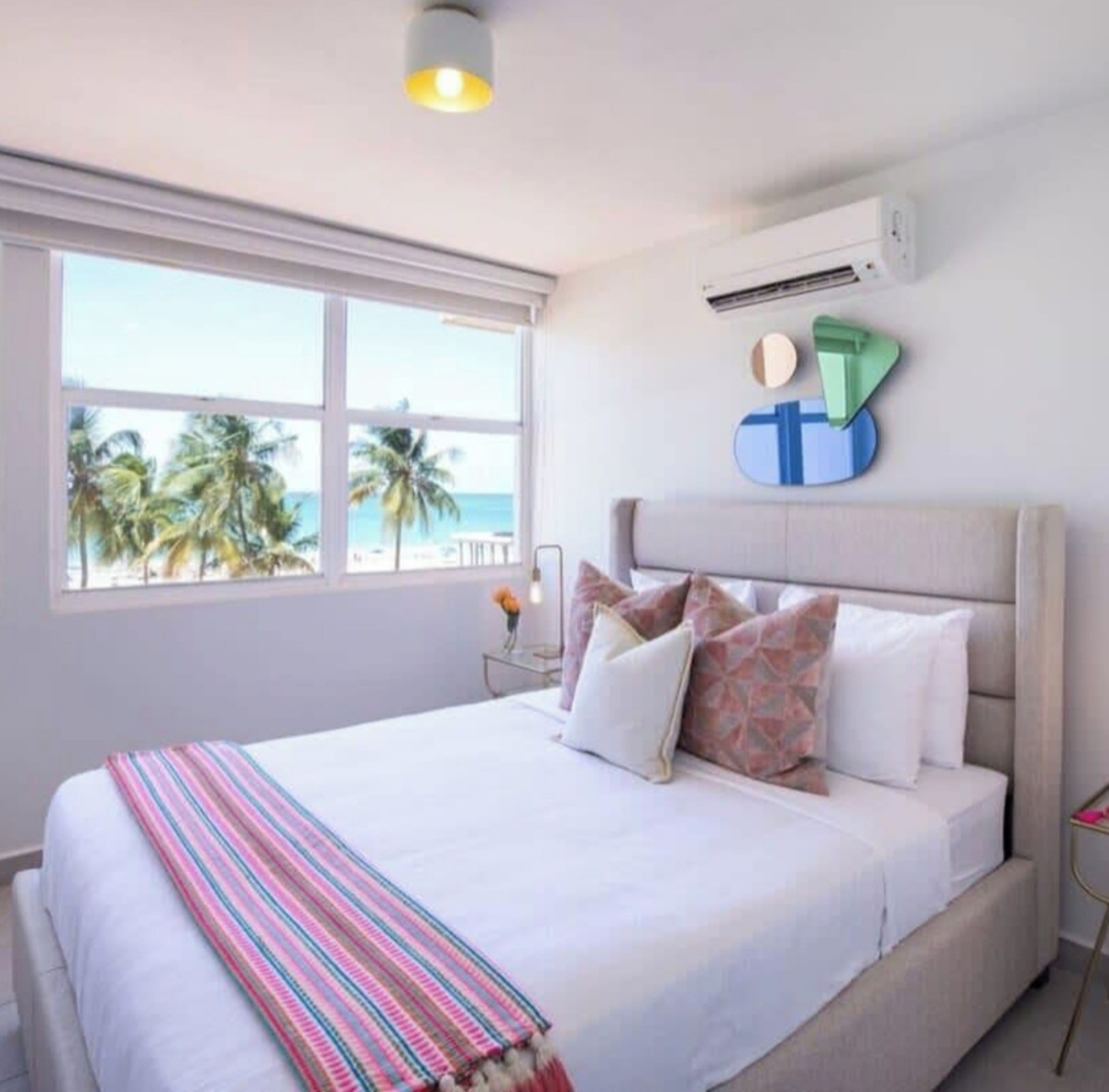 Un dormitorio con un ventanal grande, una cama con ropa de cama blanca y una manta a rayas.