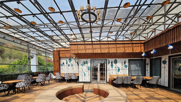 A modern indoor seating area with a central circular seating arrangement, wooden flooring, and a glass ceiling with hanging lights. - The Blu Pearl (Gwalior)