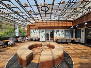 Un área de estar interior moderna con una disposición circular central, suelo de madera y techo de cristal con luces colgantes.