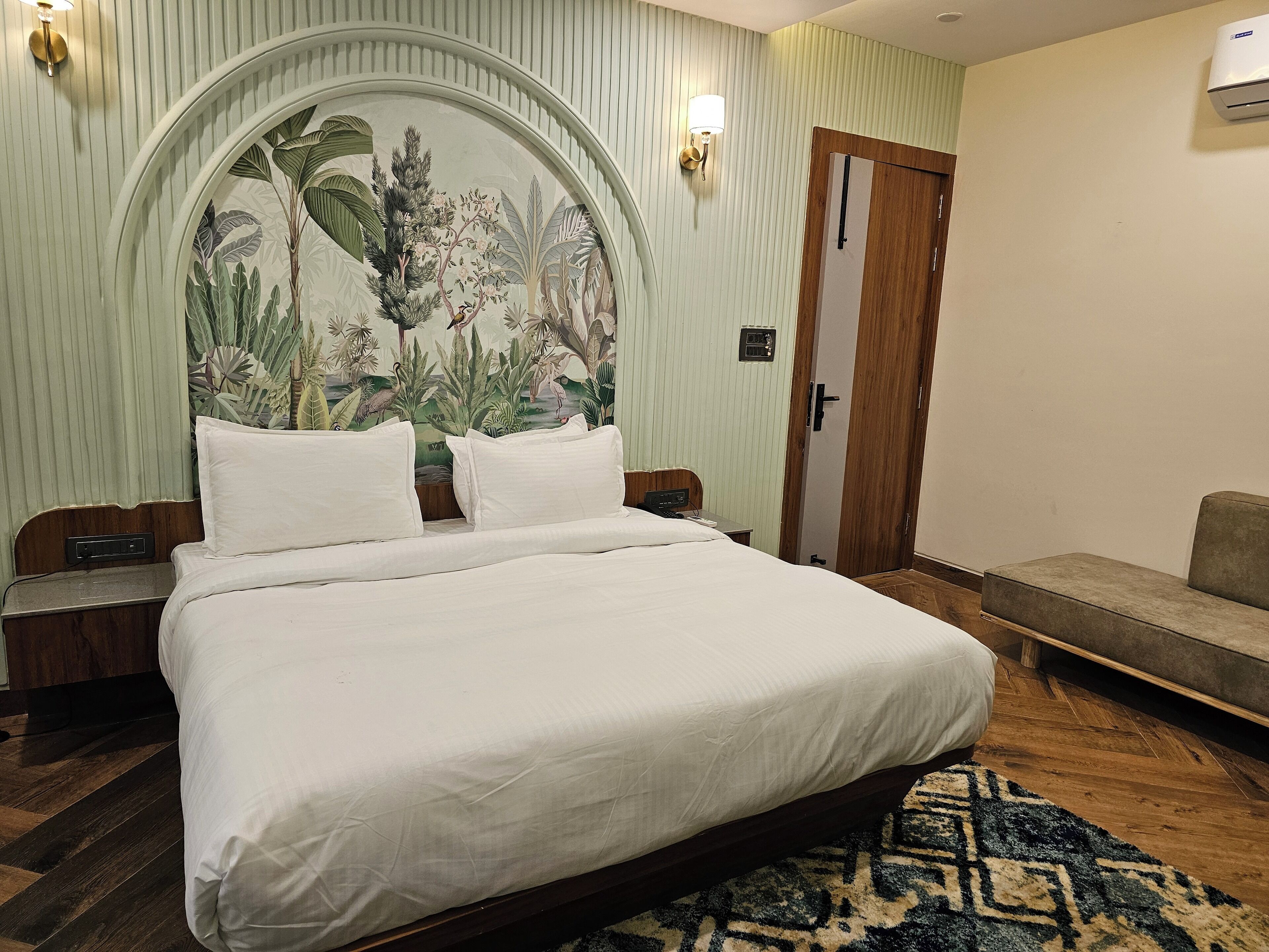 Een opgemaakt bed met witte lakens in een kamer met een houten hoofdeinde, een behang met patroon, een houten deur en een kleine bank.