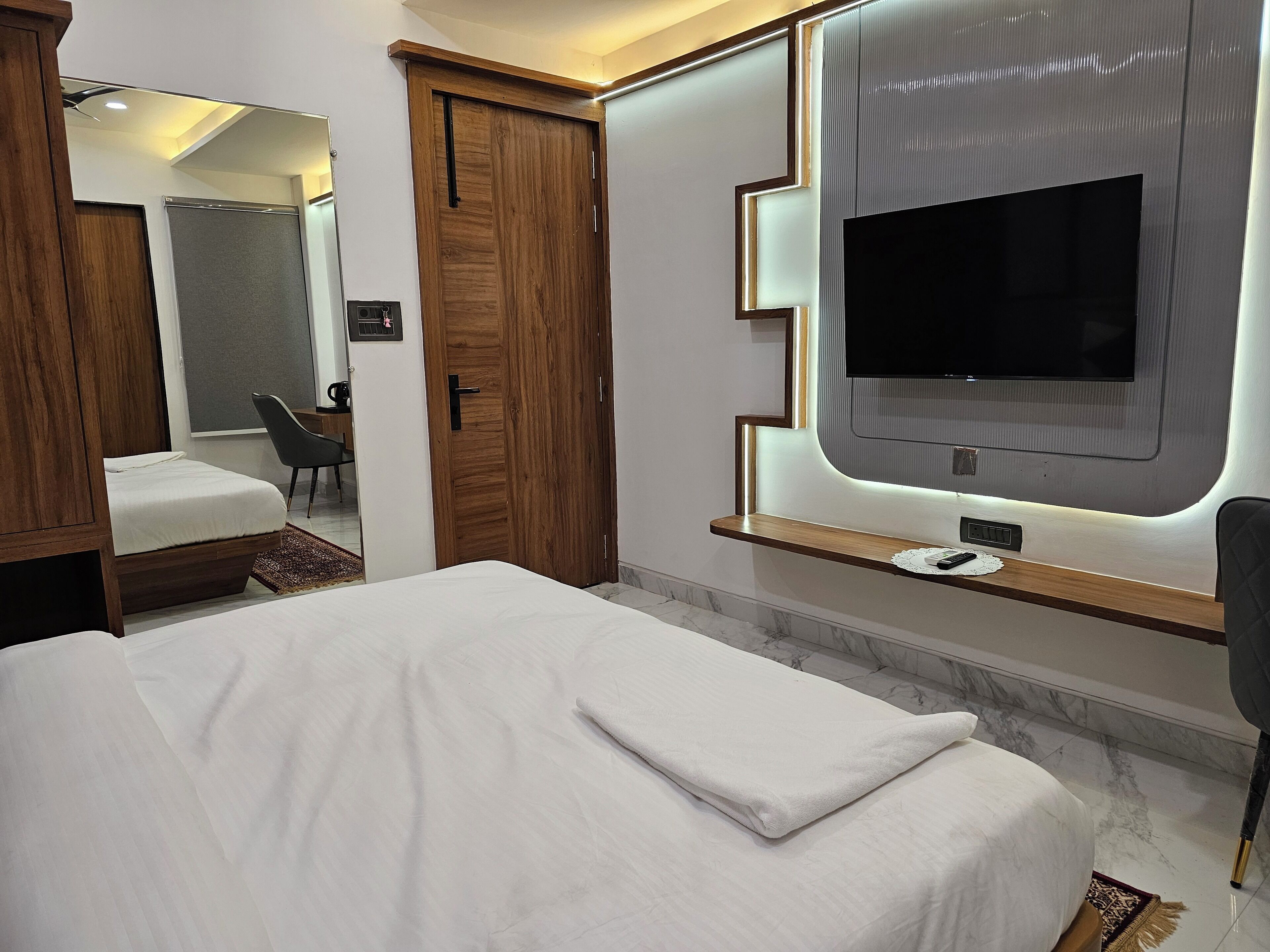 A hotel room with a bed, a television mounted on the wall, a wooden wardrobe, and a mirror.