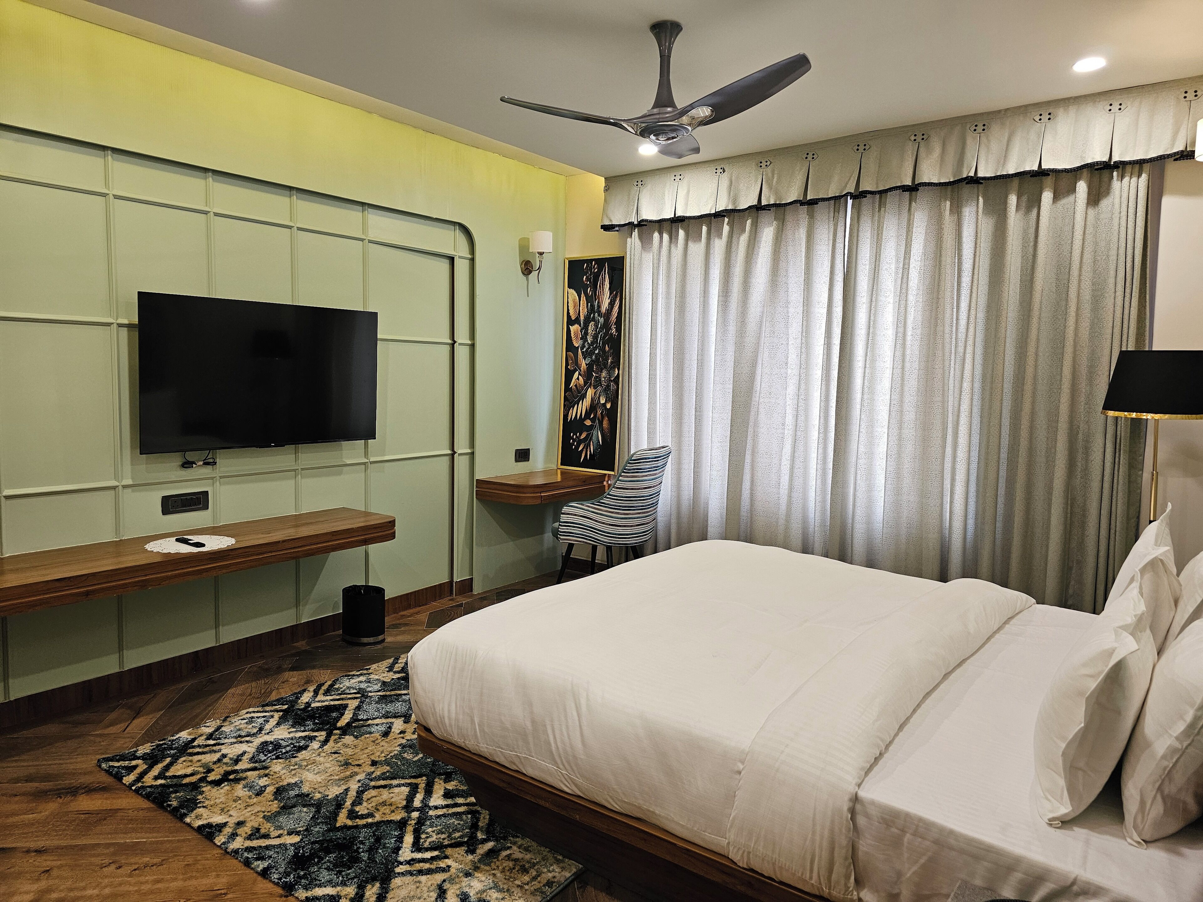 Une chambre d’hôtel avec un lit, un téléviseur fixé au mur, un ventilateur de plafond, une fenêtre avec des rideaux et une petite table avec une chaise.