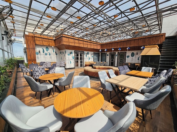 An outdoor seating area with round tables and cushioned chairs, a wooden ceiling, and decorative wall art. - The Blu Pearl (Gwalior)