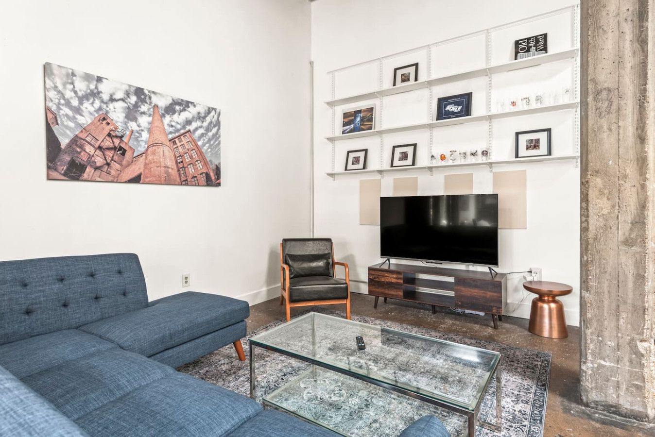 Un salon avec un canapé bleu, une table basse en verre, un fauteuil en cuir, un téléviseur à écran plat et une bibliothèque avec des photos encadrées.