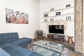 A living room with a blue sofa, a glass coffee table, a leather chair, a flat-screen TV, and a bookshelf with framed pictures. - Pet Friendly | King Bed | Beltline Access (Atlanta)
