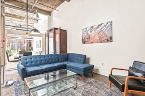 A living room with a blue sectional sofa, a glass coffee table, and a painting of buildings on the wall. - Pet Friendly | King Bed | Beltline Access (Atlanta)