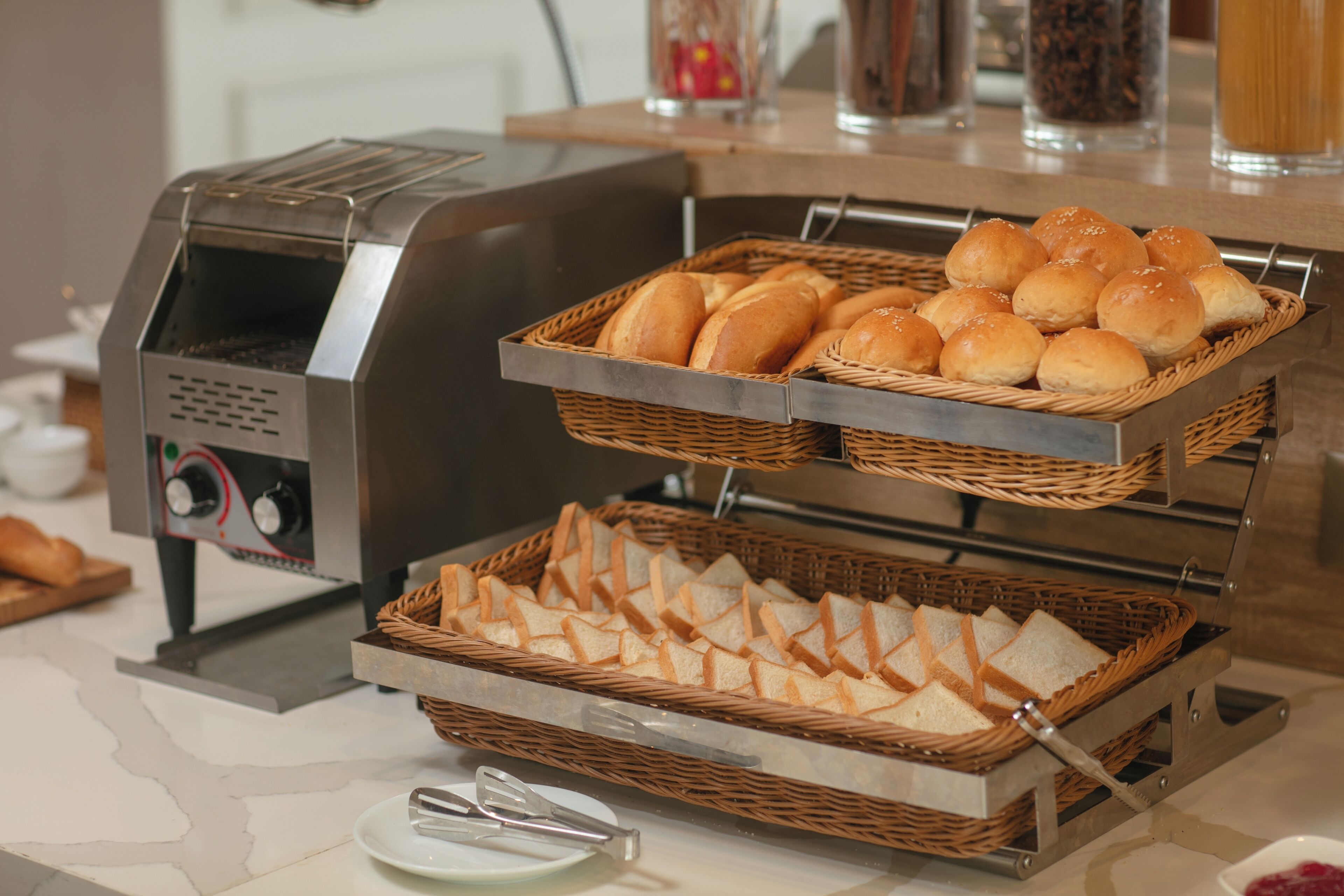 A buffet setup with a toaster oven, baskets of bread rolls, and sliced bread.