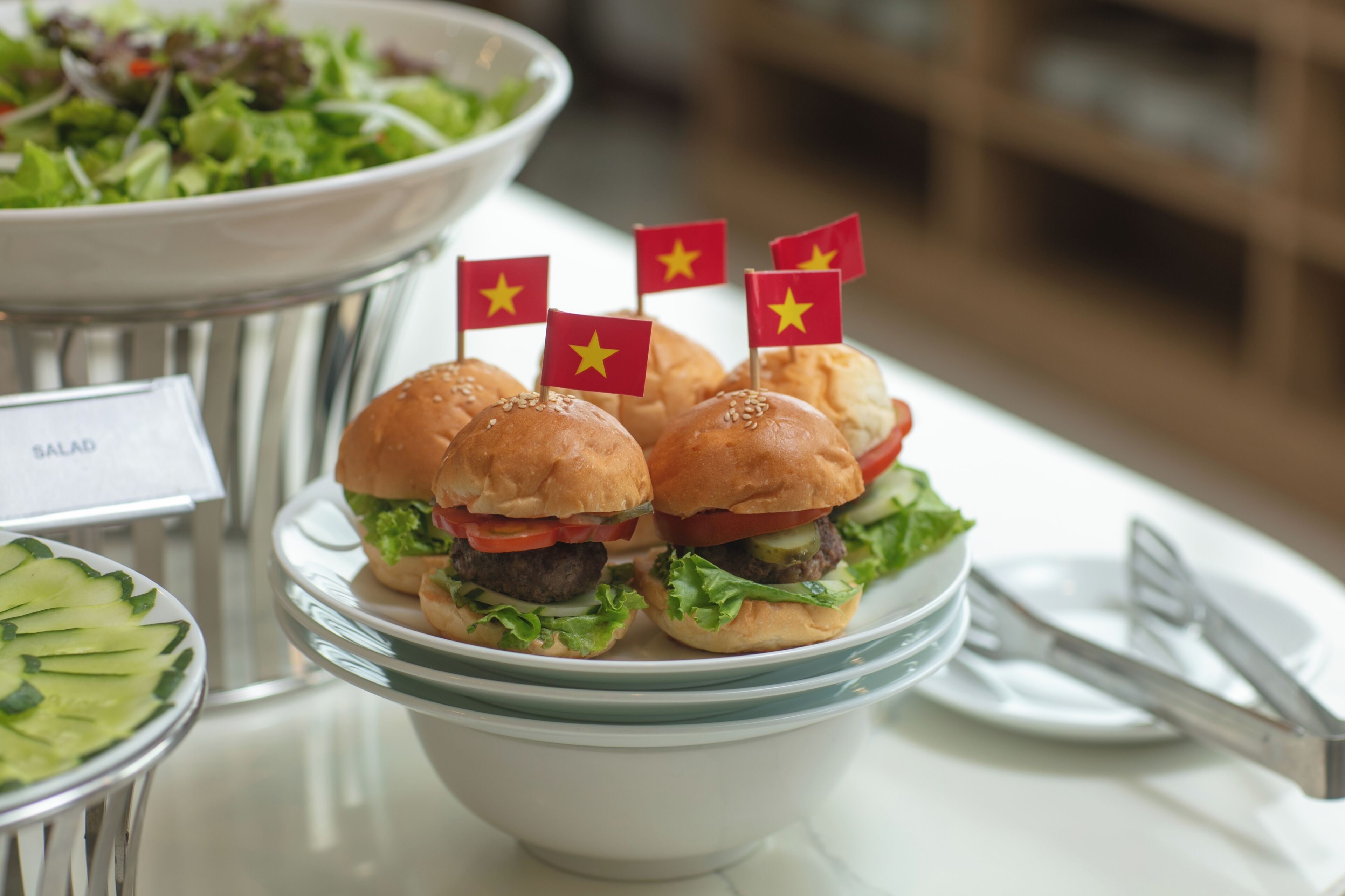 Un bol rempli de mini-burgers décorés de drapeaux vietnamiens, une salade et un panneau indiquant qu’une option salade est disponible.